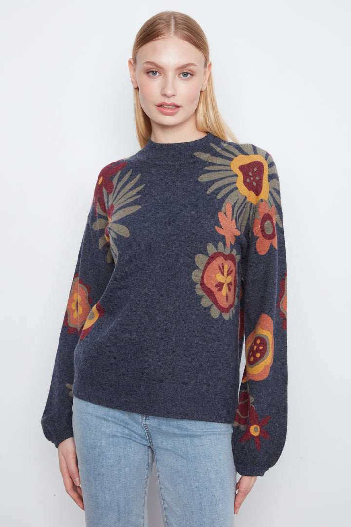 A woman modeling a dark navy blue knit sweater with a colorful floral and botanical pattern in orange, red, yellow, and beige, paired with light blue jeans against a plain white studio background.