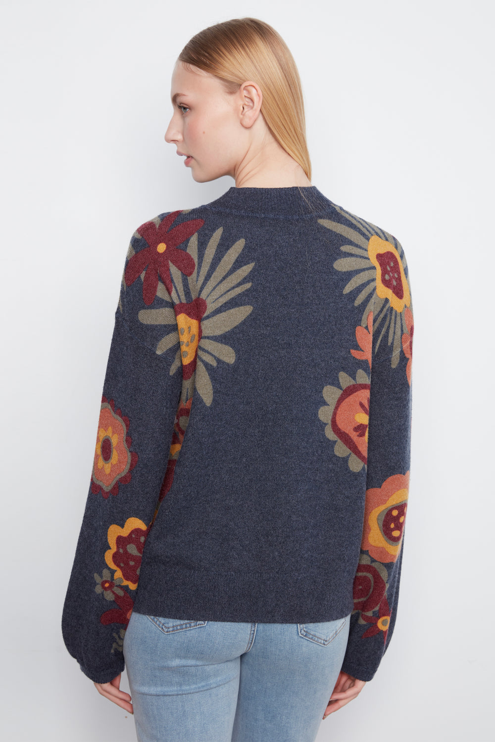 A woman shown from the back wearing a dark navy blue knit sweater with a large floral pattern in red, yellow, and beige, paired with light blue jeans against a plain white background.