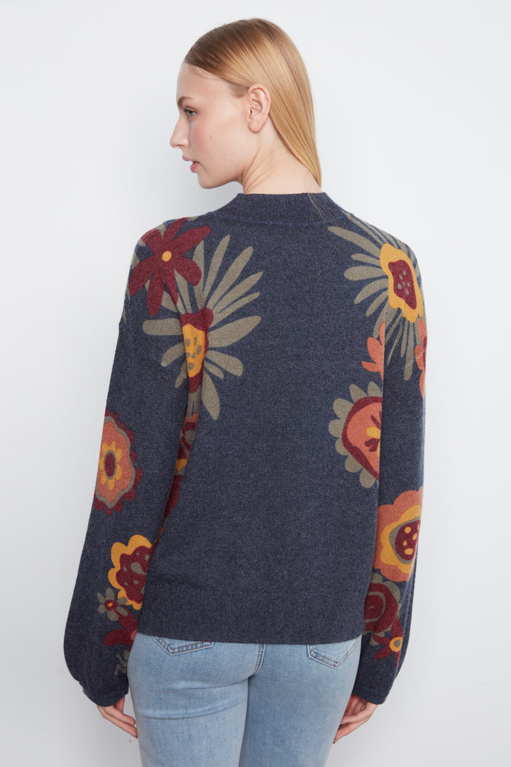 A woman shown from the back wearing a dark navy blue knit sweater with a large floral pattern in red, yellow, and beige, paired with light blue jeans against a plain white background.