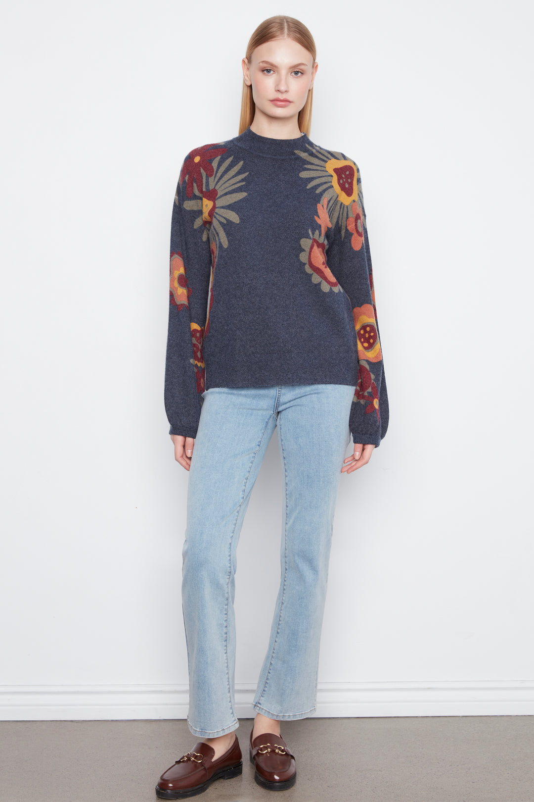 A woman stands against a plain white wall wearing a dark navy floral knit sweater, light blue jeans, and dark brown loafers.