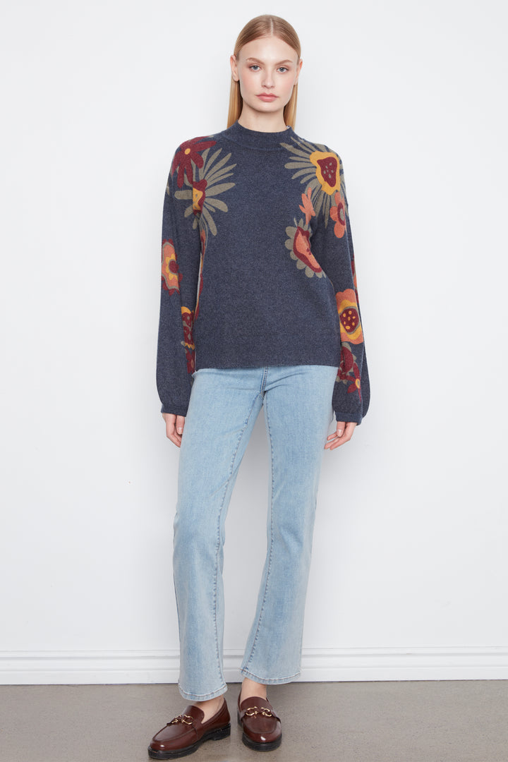 A woman stands against a plain white wall wearing a dark navy floral knit sweater, light blue jeans, and dark brown loafers.