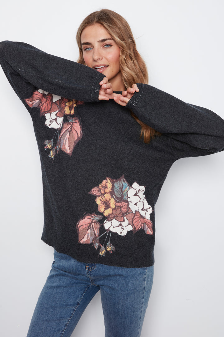 A woman wearing a dark charcoal gray knit sweater with a large multicolor floral print on the front, paired with blue jeans, posing against a light gray background.