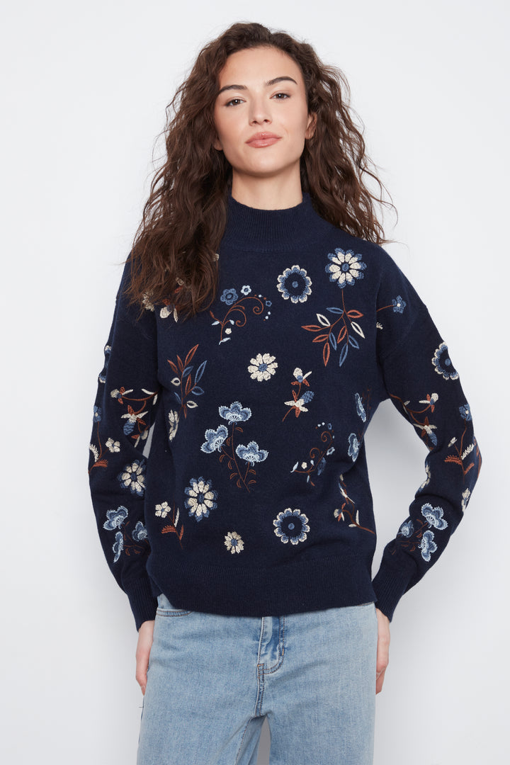 A woman wearing a navy blue mock-neck knit sweater with a colorful floral pattern, paired with light-wash blue jeans, against a plain light gray background.