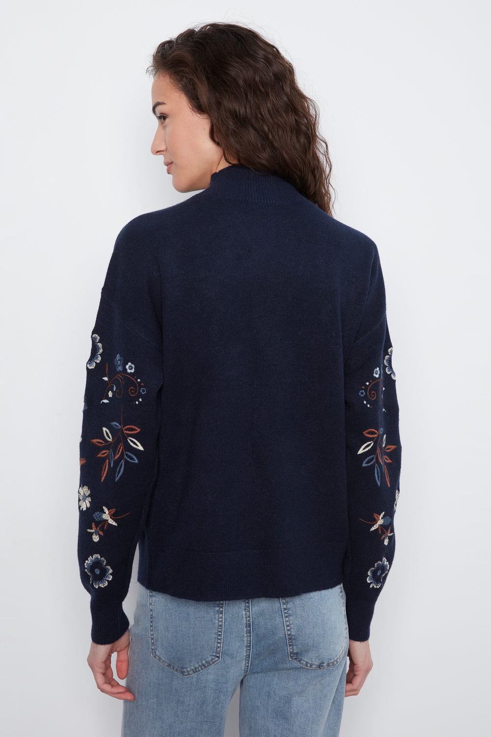 A woman shown from the back wearing a navy blue knit cardigan with colorful floral and bird embroidery on the sleeves, paired with light blue jeans, against a plain white background.