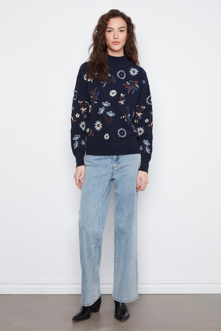 A woman stands against a white wall wearing a navy blue mock-neck knit sweater with a white, blue, and orange floral pattern, paired with light-wash wide-leg jeans and black ankle boots.