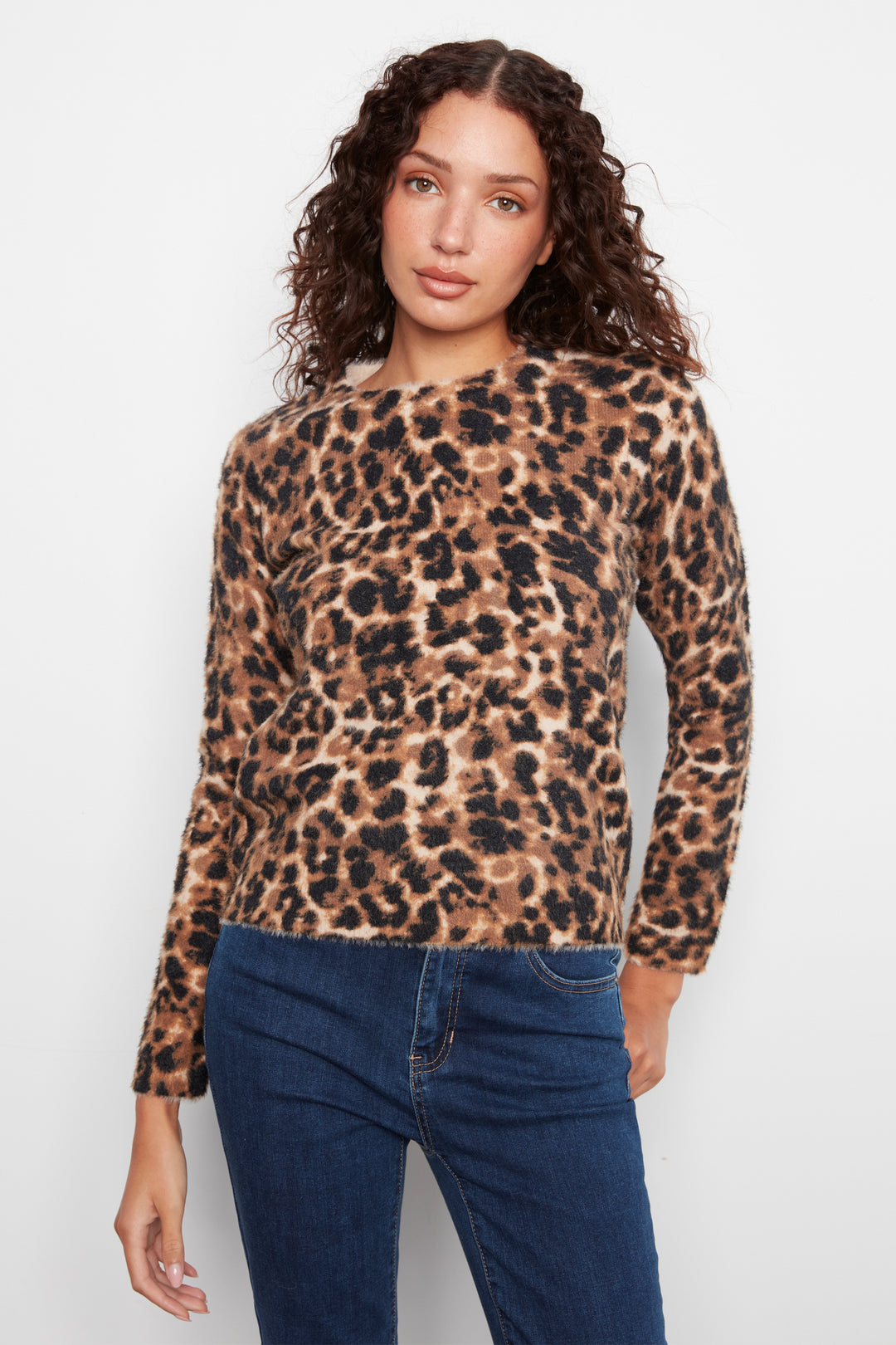 A woman modeling a long-sleeve leopard print knit top with dark blue jeans, photographed against a plain light gray studio background.