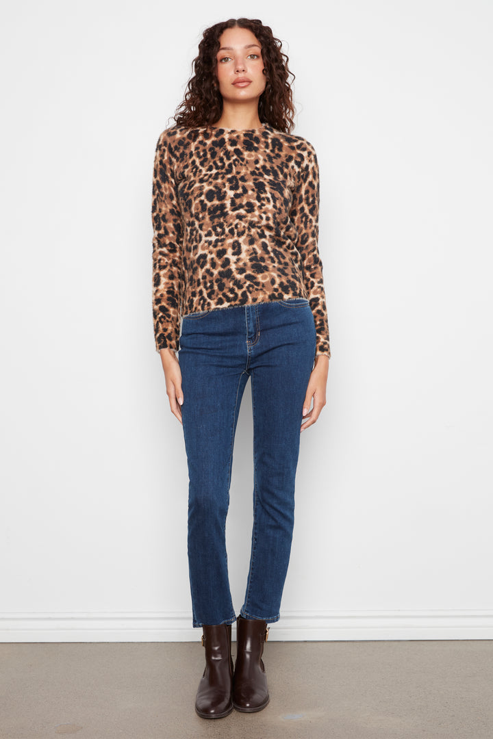 A woman stands against a plain white wall wearing a leopard-print long-sleeve top, dark blue jeans, and dark brown ankle boots.