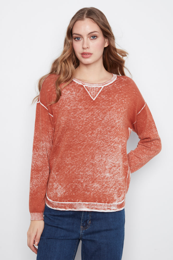 A woman modeling a long-sleeve crewneck sweater in a washed terracotta orange color with white contrast stitching and a white geometric V-neck detail, paired with blue jeans against a light gray background.