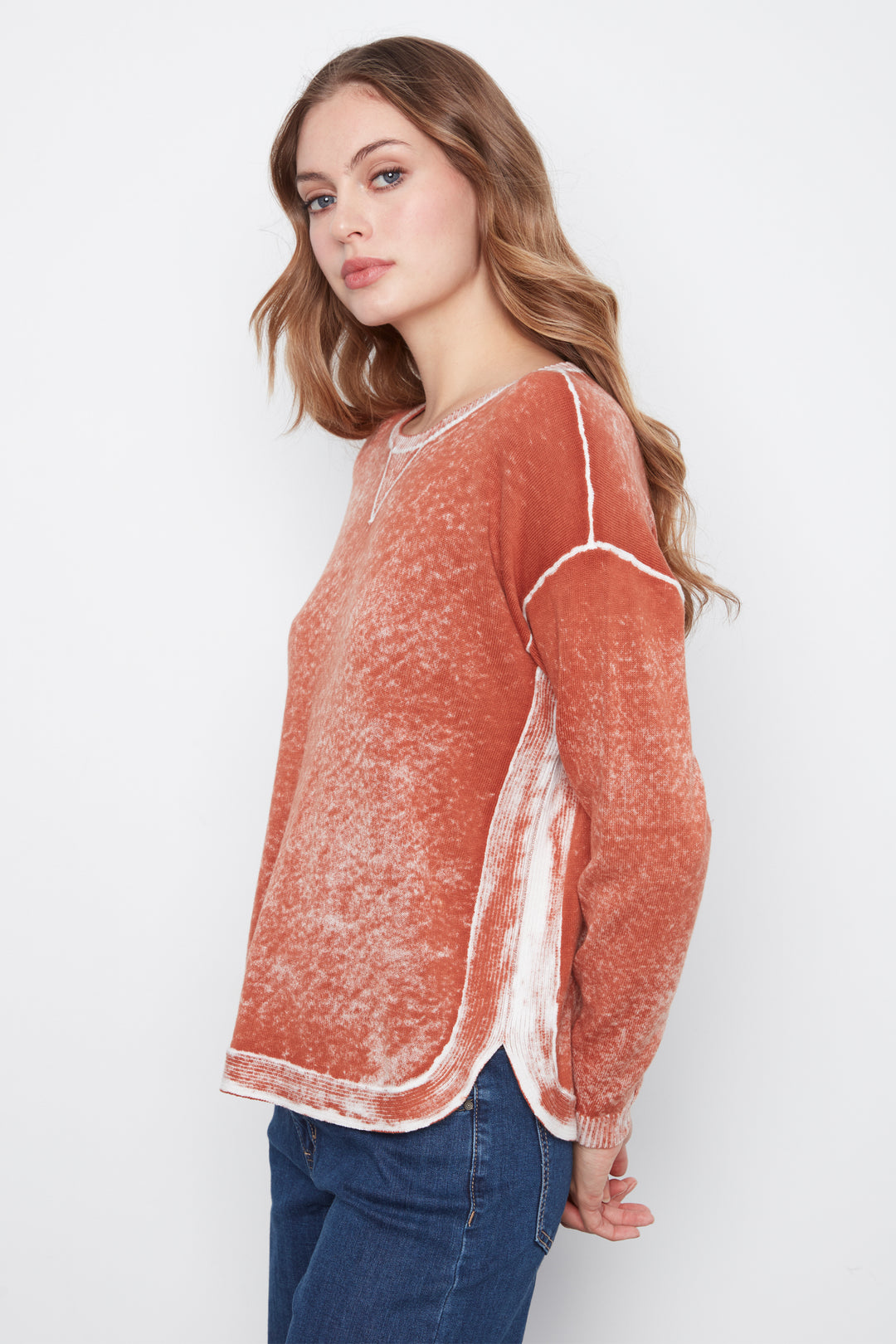 A woman wearing a rust-orange long-sleeve knit sweater with white contrast stitching and side panels, paired with blue jeans, photographed against a light gray studio background.