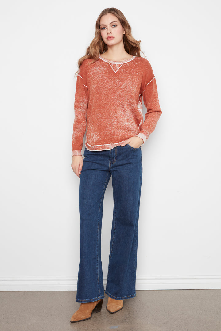 A woman modeling a rust-orange textured knit sweater with white contrast trim and blue wide-leg jeans, standing against a plain white wall.