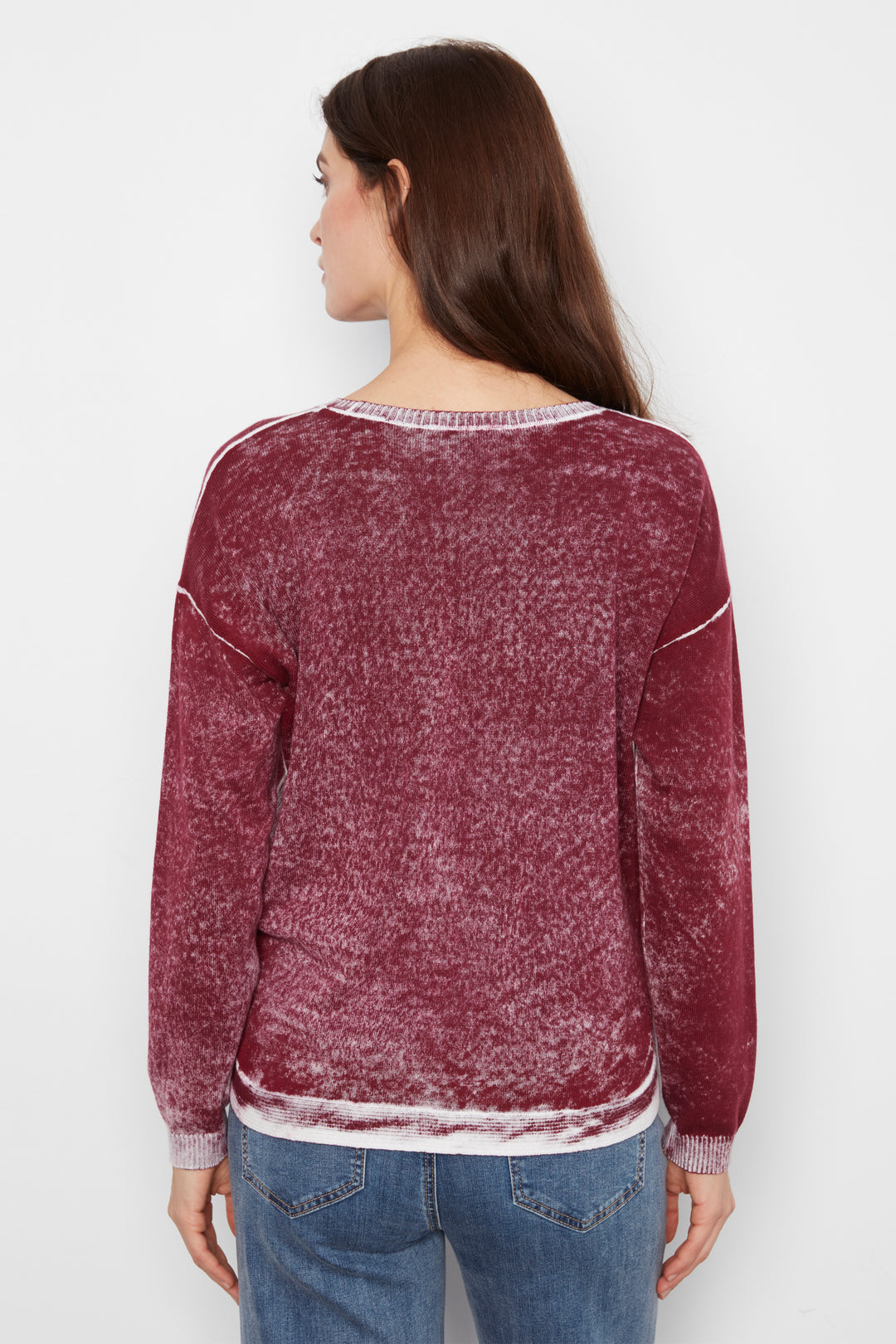 A woman viewed from the back wearing a marled, washed burgundy long-sleeve sweater with white contrast stitching and a white hem, paired with blue jeans against a plain white background.