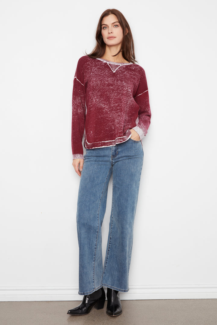 A woman stands against a plain white wall wearing a maroon long-sleeve knit sweater with white contrast stitching and a V-neckline, paired with wide-leg blue jeans and black ankle boots.