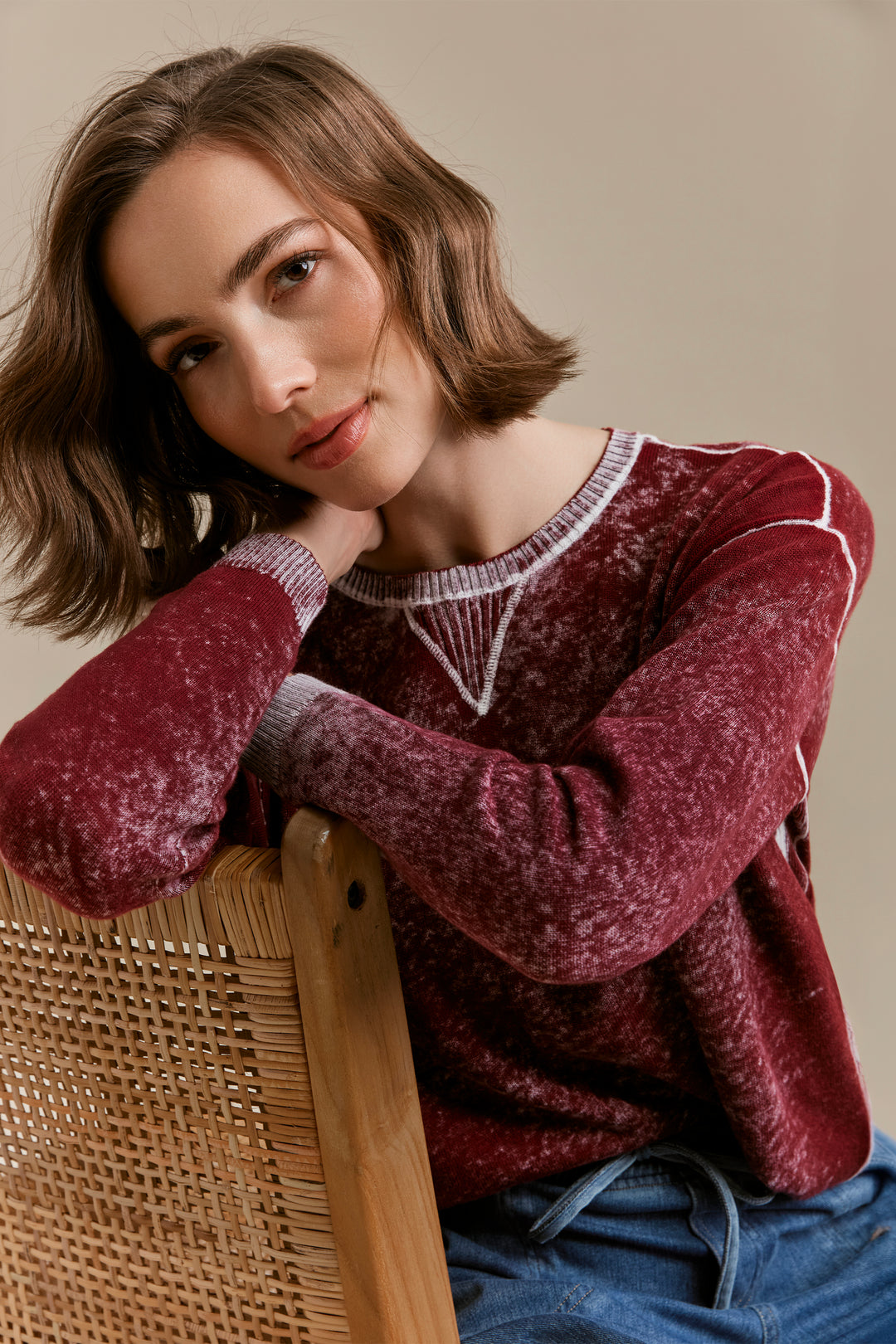 A woman with short brown hair leans on a woven chair, wearing a maroon textured knit sweater with white geometric accents on the chest and cuffs, paired with blue jeans against a warm beige background.