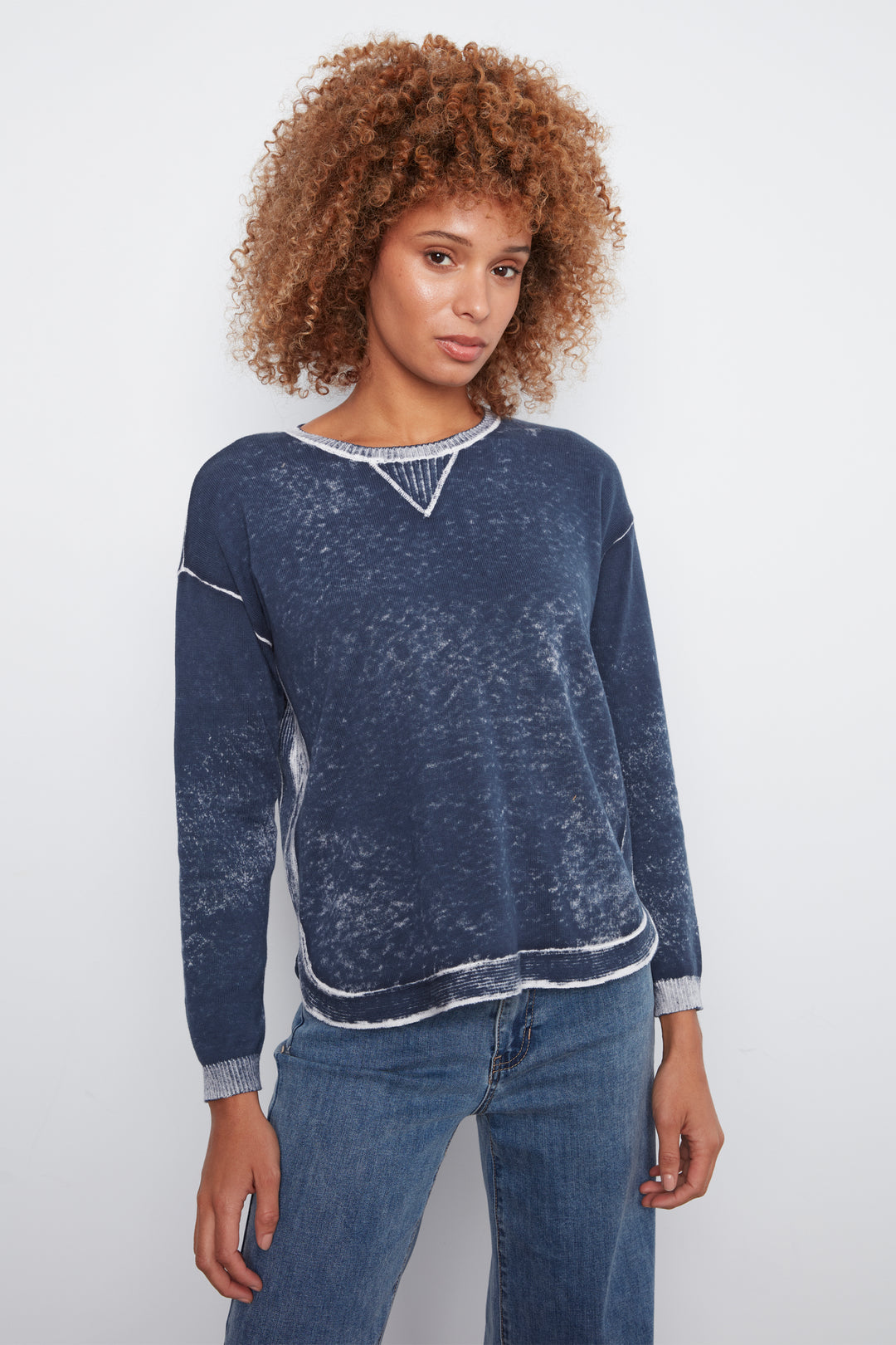 A woman with curly light brown hair wearing a dark blue acid-wash long-sleeve sweater with white contrast stitching and blue jeans, standing against a light gray studio background.