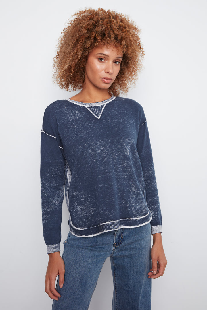 A woman with curly light brown hair wearing a dark blue acid-wash long-sleeve sweater with white contrast stitching and blue jeans, standing against a light gray studio background.