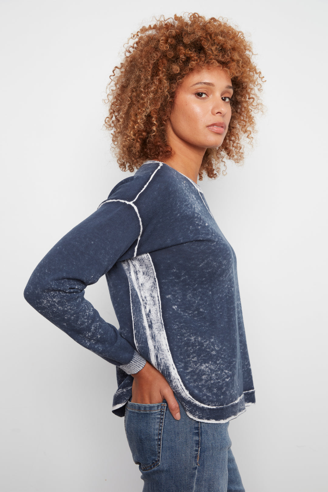 A woman with curly brown hair wearing a dark blue acid-wash long-sleeve top with white contrast stitching and side pockets, paired with blue jeans, photographed against a light gray studio background.