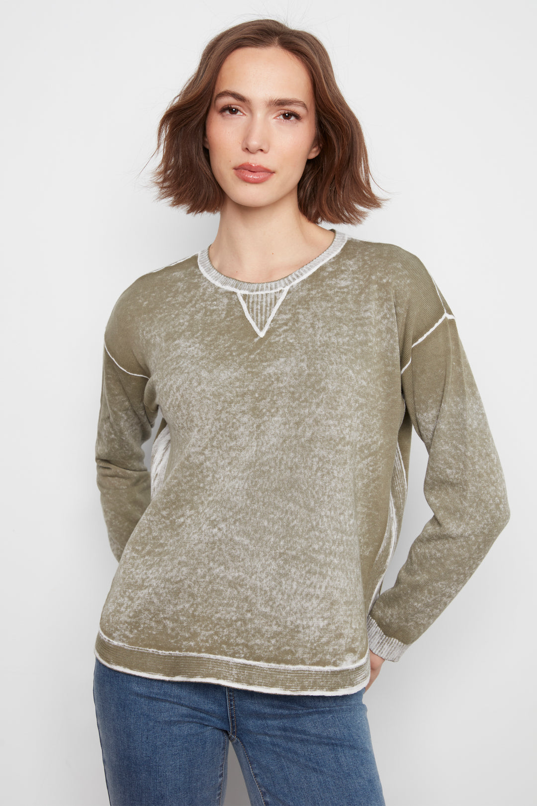 A woman modeling a long-sleeve crewneck sweater in a heathered olive green color with white contrast stitching and a textured knit V-neck detail, paired with blue jeans against a light gray background.