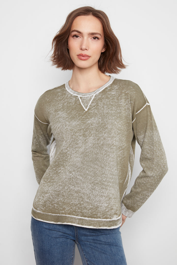 A woman modeling a long-sleeve crewneck sweater in a heathered olive green color with white contrast stitching and a textured knit V-neck detail, paired with blue jeans against a light gray background.