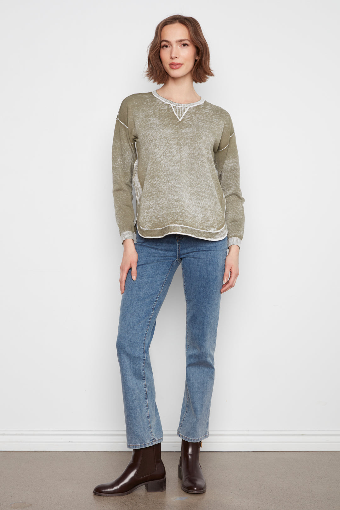 A woman modeling a heathered sage green knit sweater with white lace trim at the neckline, paired with blue jeans and dark brown leather ankle boots, standing against a plain white wall.