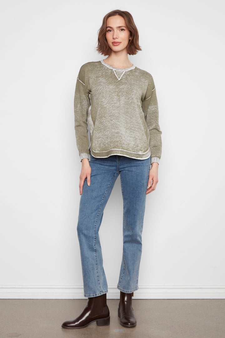 A woman modeling a heathered sage green knit sweater with white lace trim at the neckline, paired with blue jeans and dark brown leather ankle boots, standing against a plain white wall.