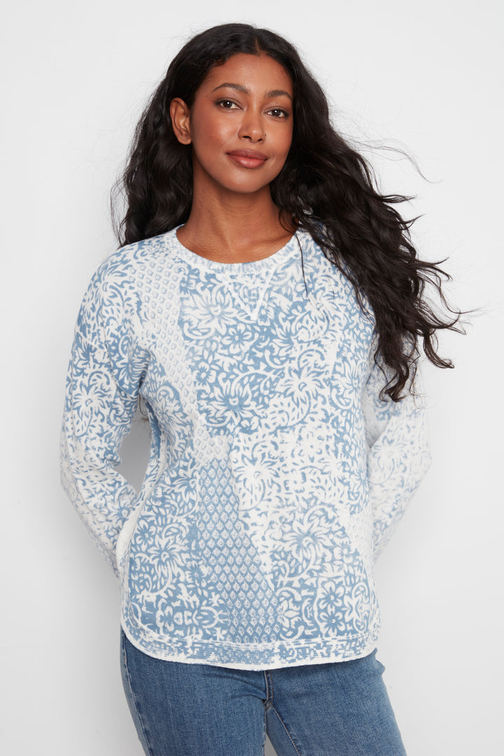 A woman modeling a light blue and white patterned knit sweater with long sleeves, paired with blue jeans, against a plain light gray background.