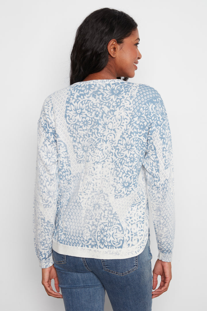 A woman shown from the back wearing a white long-sleeve top with a light blue ornate floral pattern, paired with blue jeans, against a plain white background.