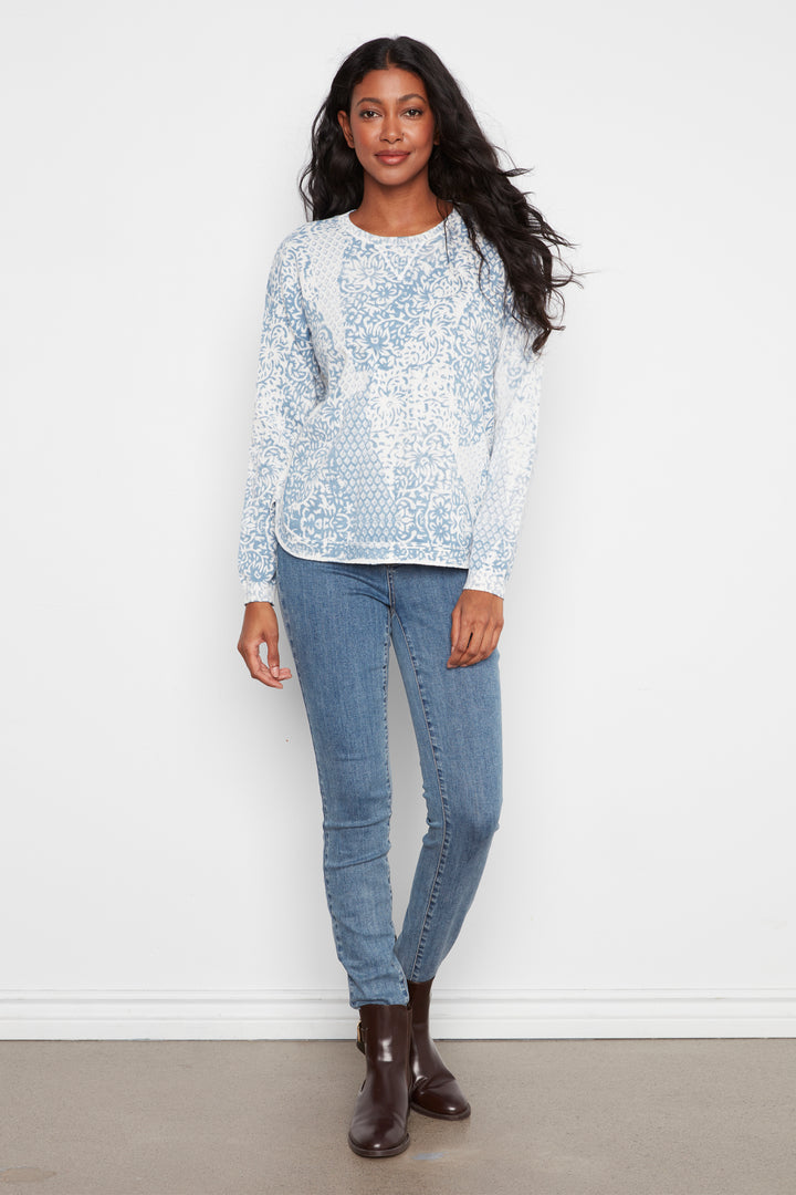 A woman modeling a light blue and white patterned long-sleeve knit top with blue skinny jeans and dark brown ankle boots, standing against a plain white wall.