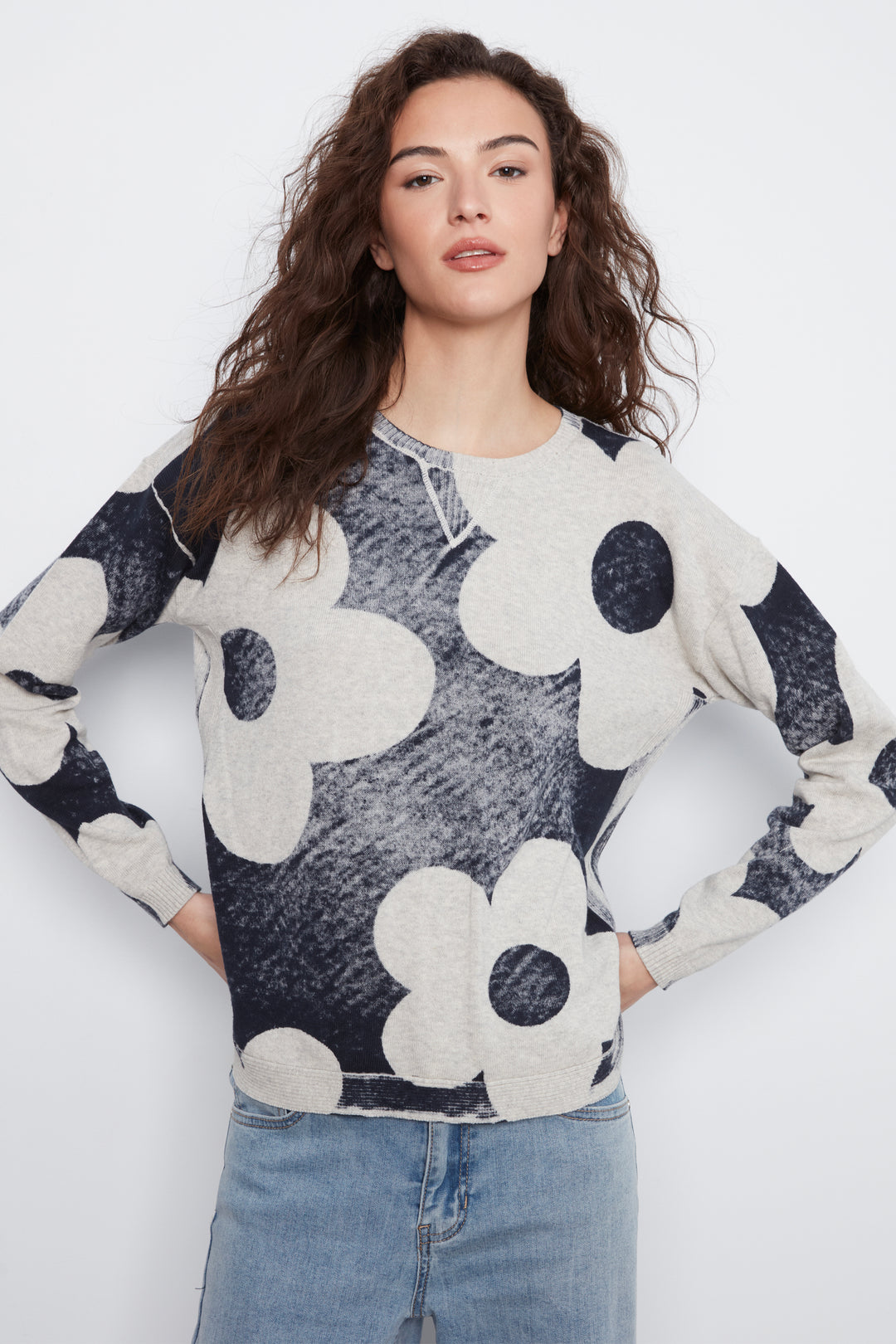 A woman modeling a long-sleeve knit top with a large-scale floral pattern in light heather gray and dark charcoal gray, paired with light-wash blue jeans against a plain white studio background.