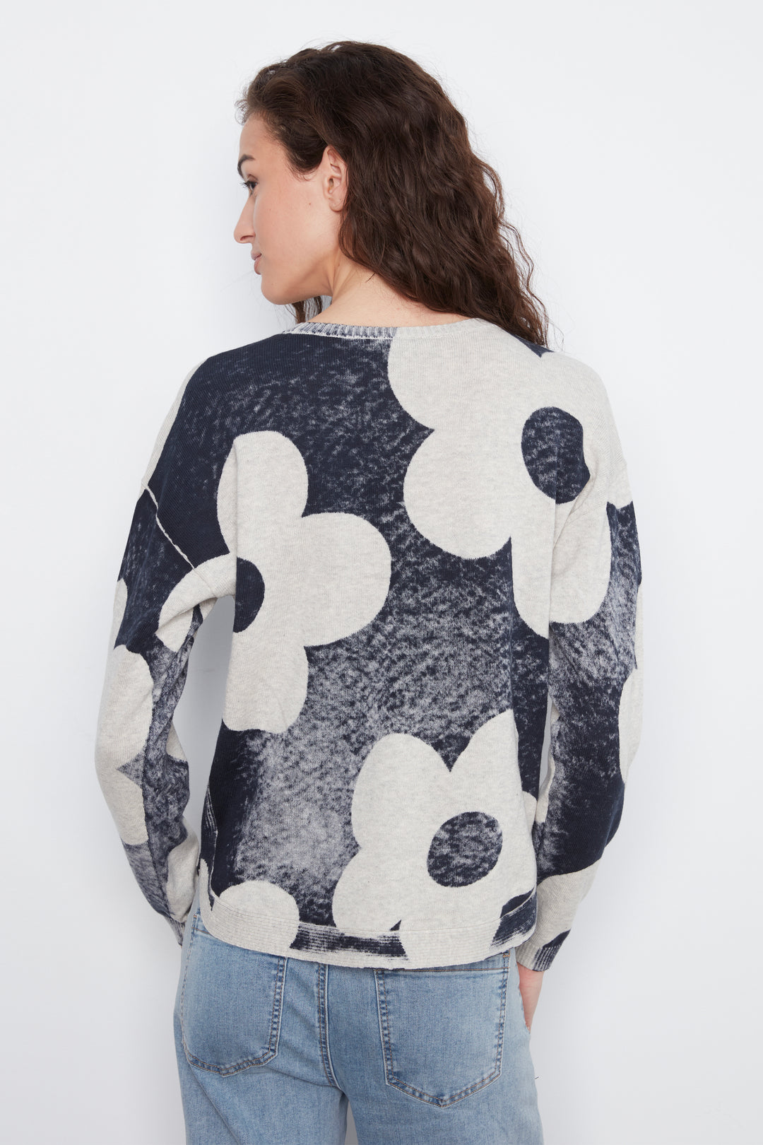 A woman viewed from the back wearing a long-sleeve knit sweater with a large-scale floral pattern in dark navy and off-white, paired with light blue jeans against a plain white background.