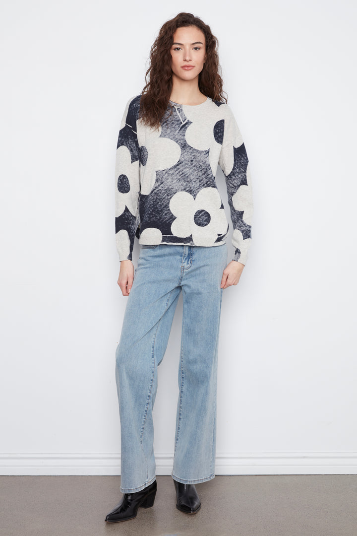 A woman modeling a long-sleeve knit sweater with a black, white, and gray floral pattern, paired with light-wash wide-leg jeans and black ankle boots, standing against a white wall.