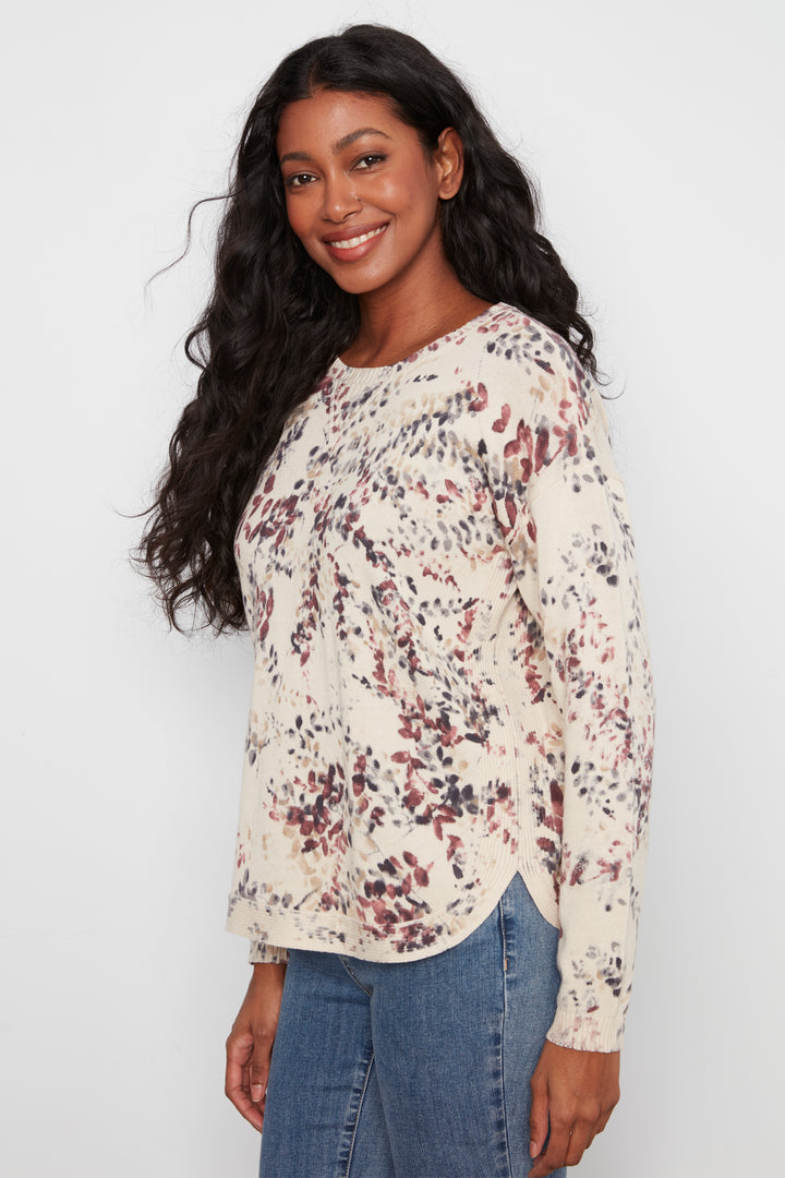A woman with long, wavy black hair is wearing a cream-colored long-sleeve top with a red and black abstract floral pattern, paired with blue jeans, against a plain light gray background.