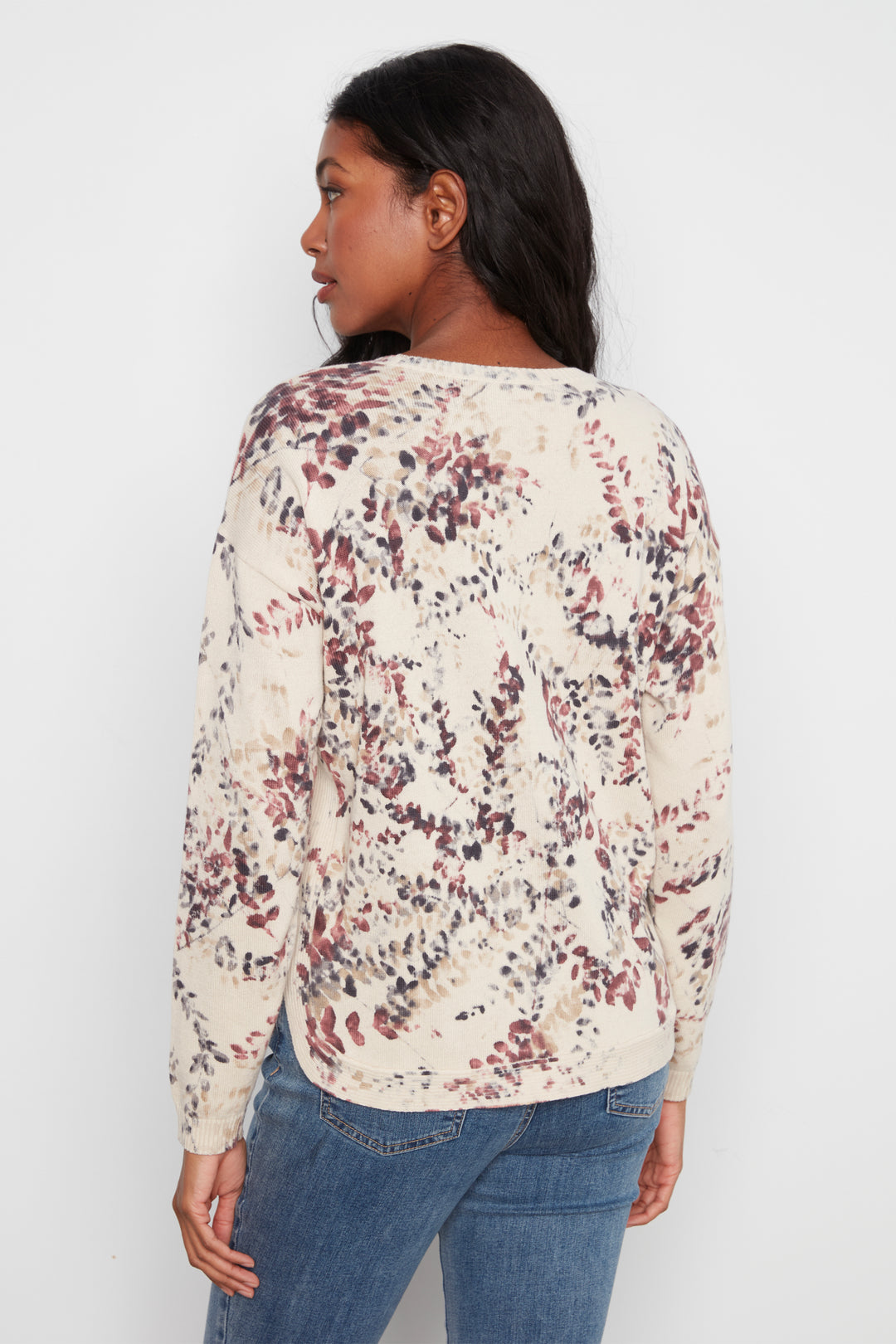 A woman shown from the back wearing a cream-colored long-sleeve top with a dense abstract floral pattern in dusty red, brown, and dark gray, paired with blue jeans against a plain white background.