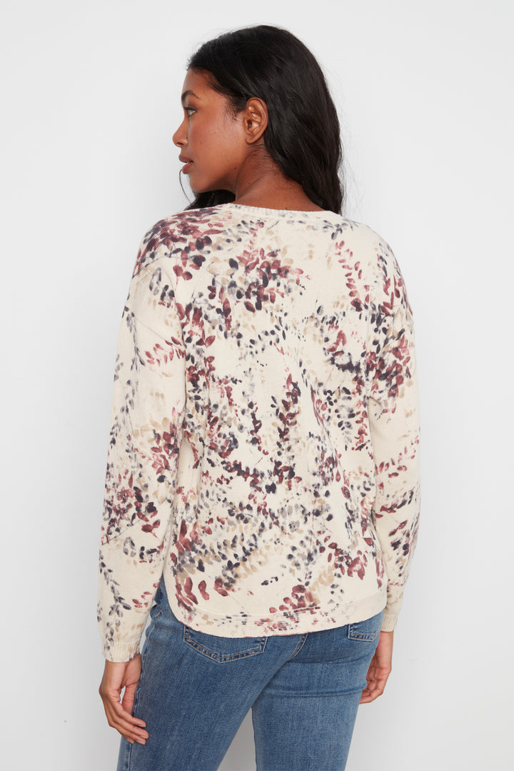 A woman shown from the back wearing a cream-colored long-sleeve top with a dense abstract floral pattern in dusty red, brown, and dark gray, paired with blue jeans against a plain white background.