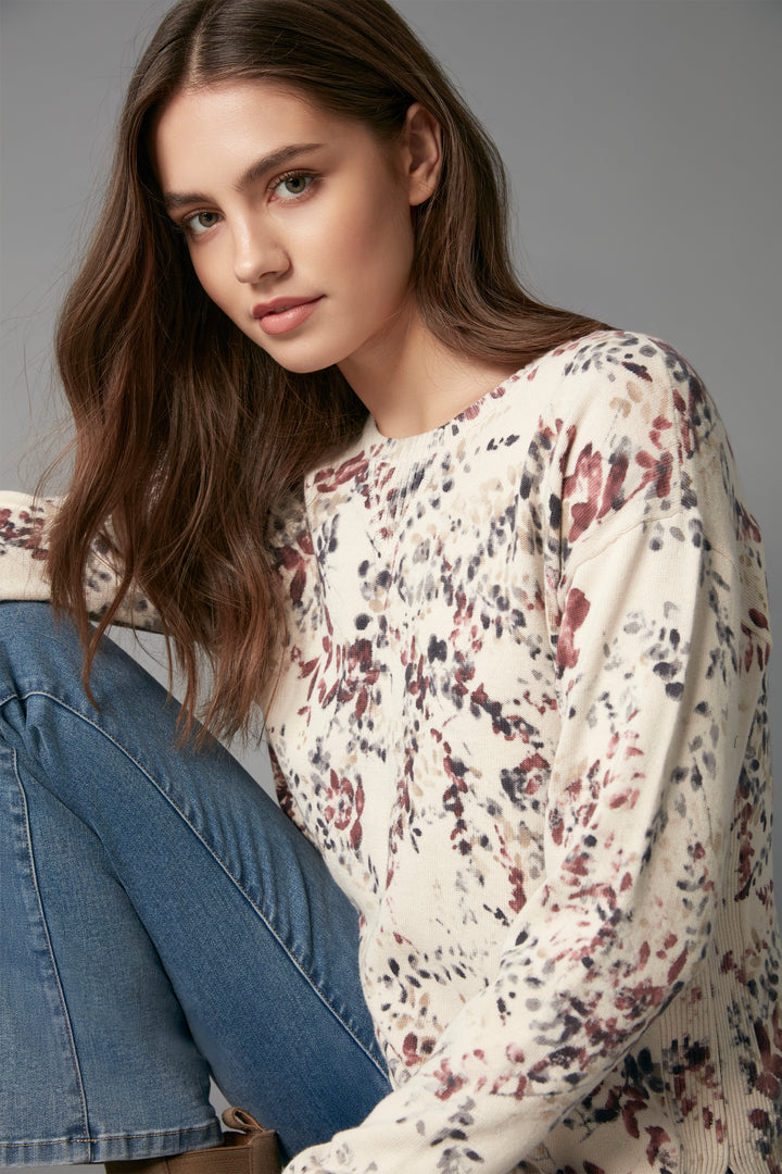 A woman wearing a cream-colored long-sleeve top with a red, black, and gray abstract floral pattern, paired with blue jeans, posed against a solid gray studio background.