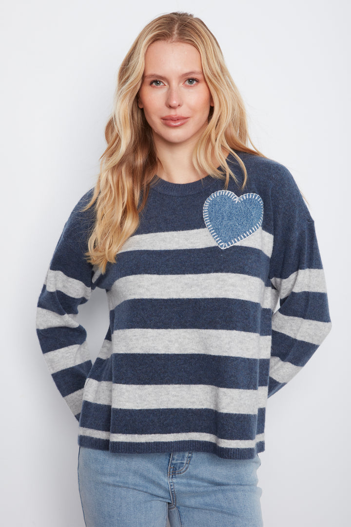 A woman with blonde hair wearing a navy and gray striped knit sweater with a blue heart patch on the chest, paired with light blue jeans, against a light gray background.