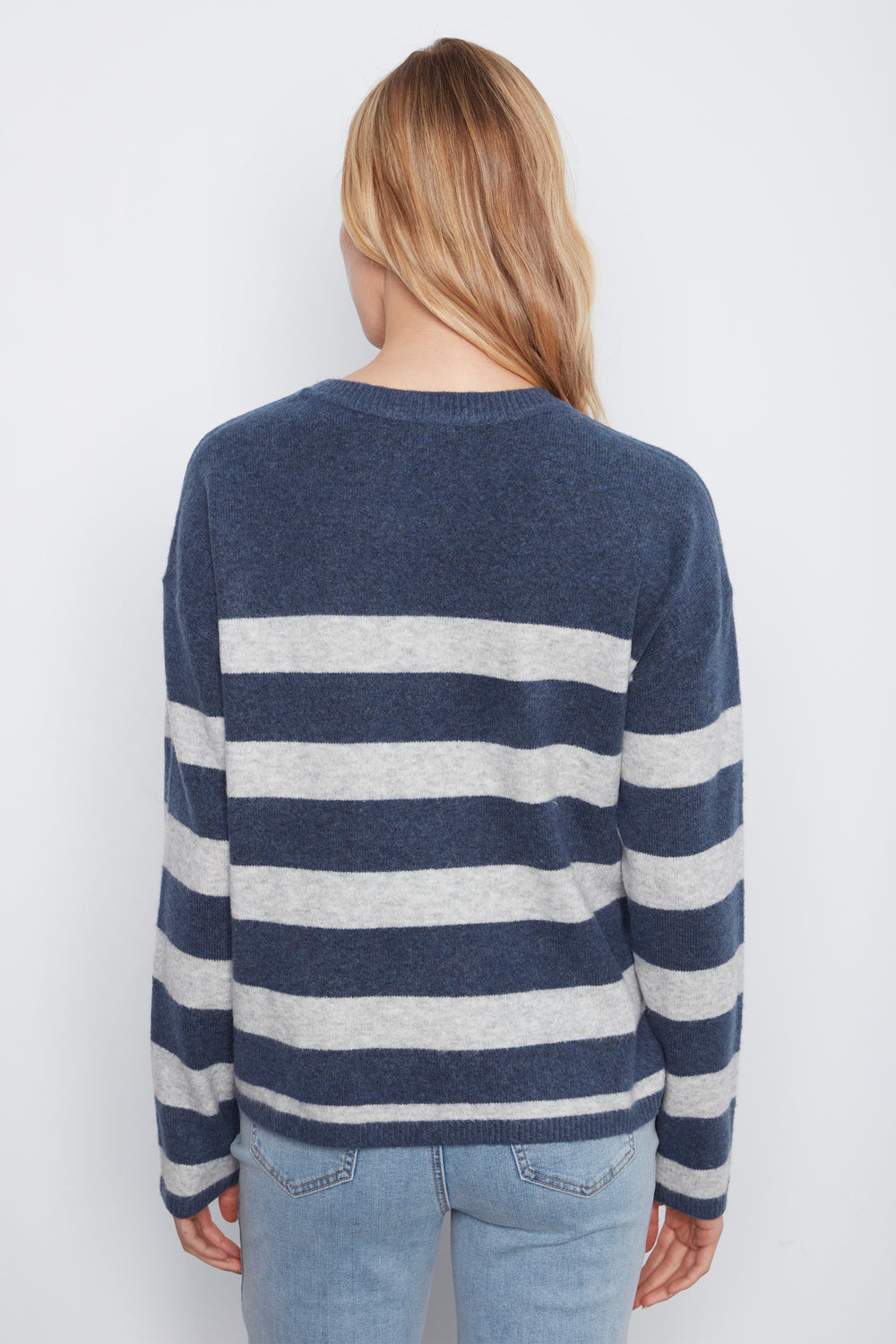 A woman viewed from the back wearing a navy and light gray striped knit sweater with light blue jeans, set against a plain light gray studio background.