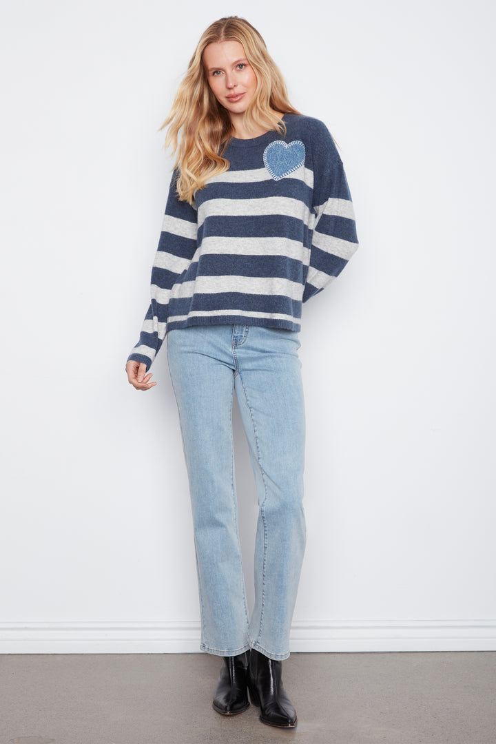 A woman modeling a navy and white striped knit sweater with a blue heart patch on the chest, paired with light blue jeans and black boots, standing against a white wall.
