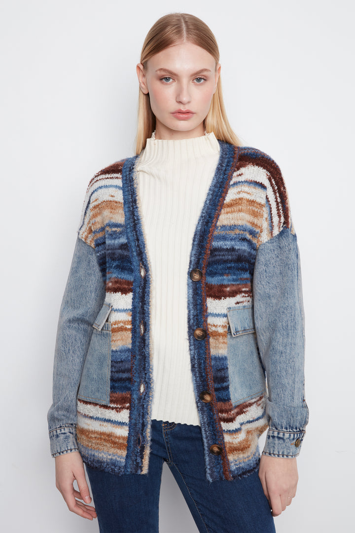 A woman modeling a chunky knit cardigan with a color-blocked pattern of blue denim, brown, beige, and white stripes, worn over a cream ribbed turtleneck and blue jeans against a light gray studio background.