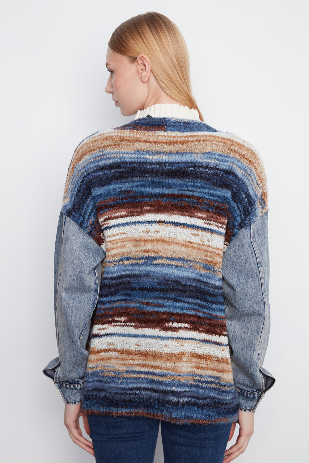 A woman shown from the back wearing a knit sweater with horizontal stripes in blue, brown, beige, and white, featuring denim sleeves, against a plain light gray background.