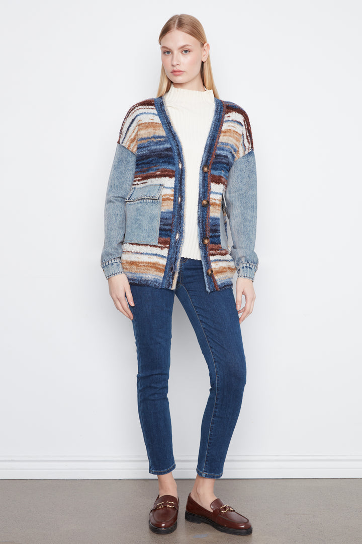 A woman modeling a mixed-media cardigan with denim sleeves and a striped knit body in shades of blue, brown, and cream, paired with dark blue jeans, a white turtleneck, and brown loafers against a white wall.