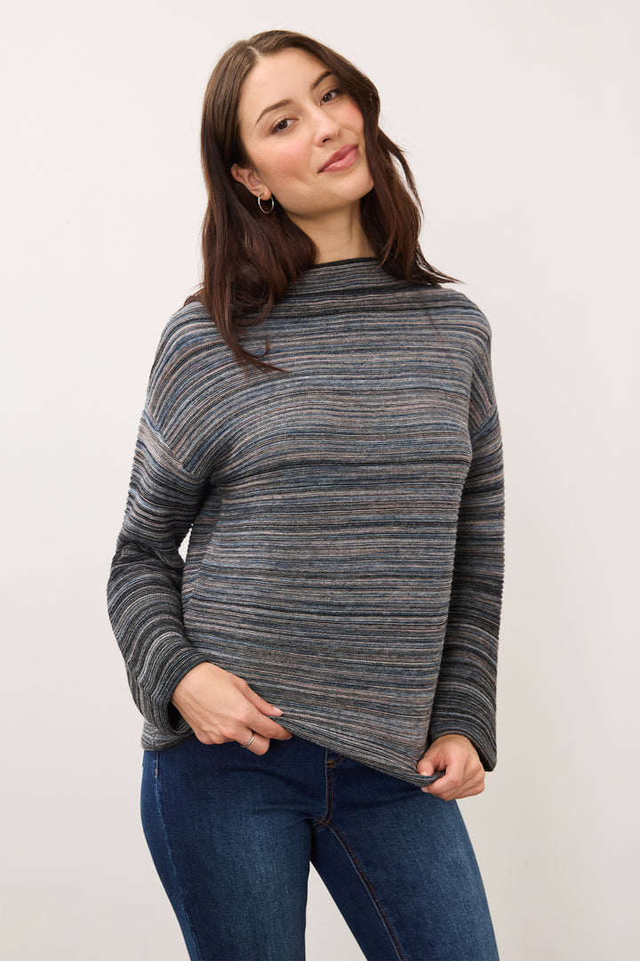 A woman wearing a gray and navy striped knit sweater with blue jeans, standing against a plain light gray background.
