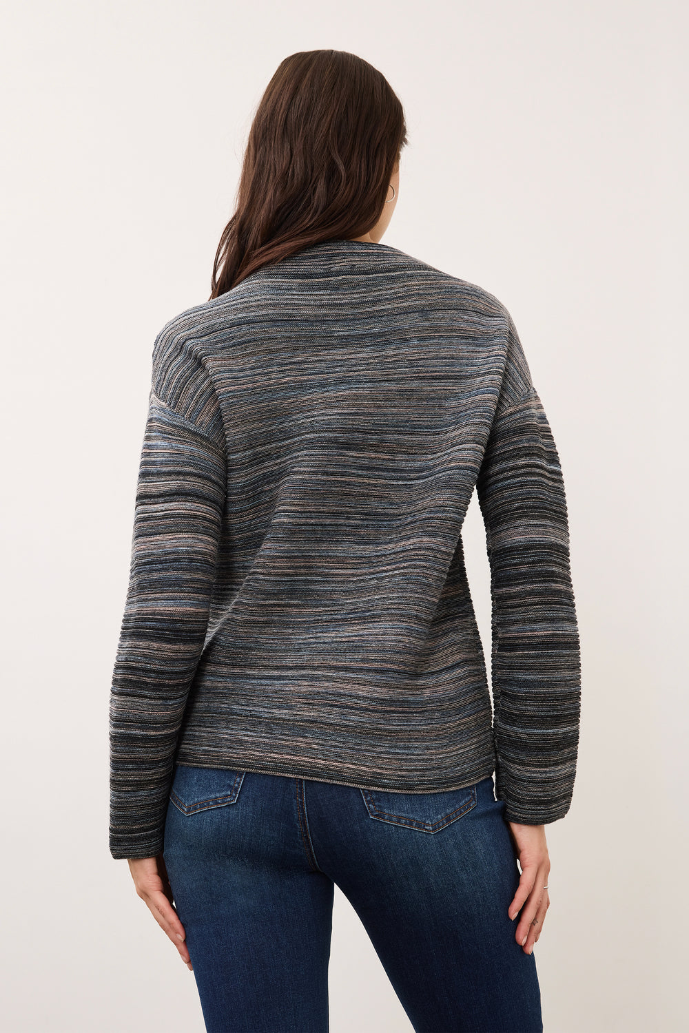 A woman viewed from the back wearing a long-sleeve knit sweater with horizontal gray and navy stripes, paired with dark blue jeans, against a plain off-white studio background.