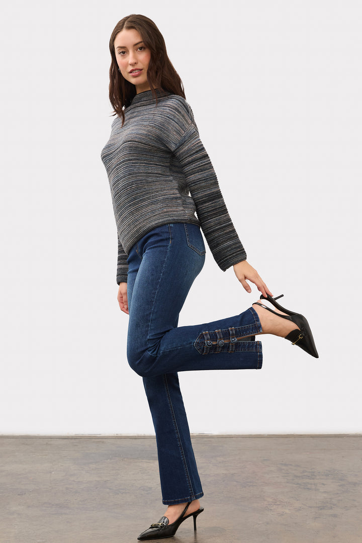 A woman modeling a gray and black striped knit mock-neck sweater, blue flare jeans with patchwork details on the back of the knees, and black pointed-toe mule heels against a plain studio background.