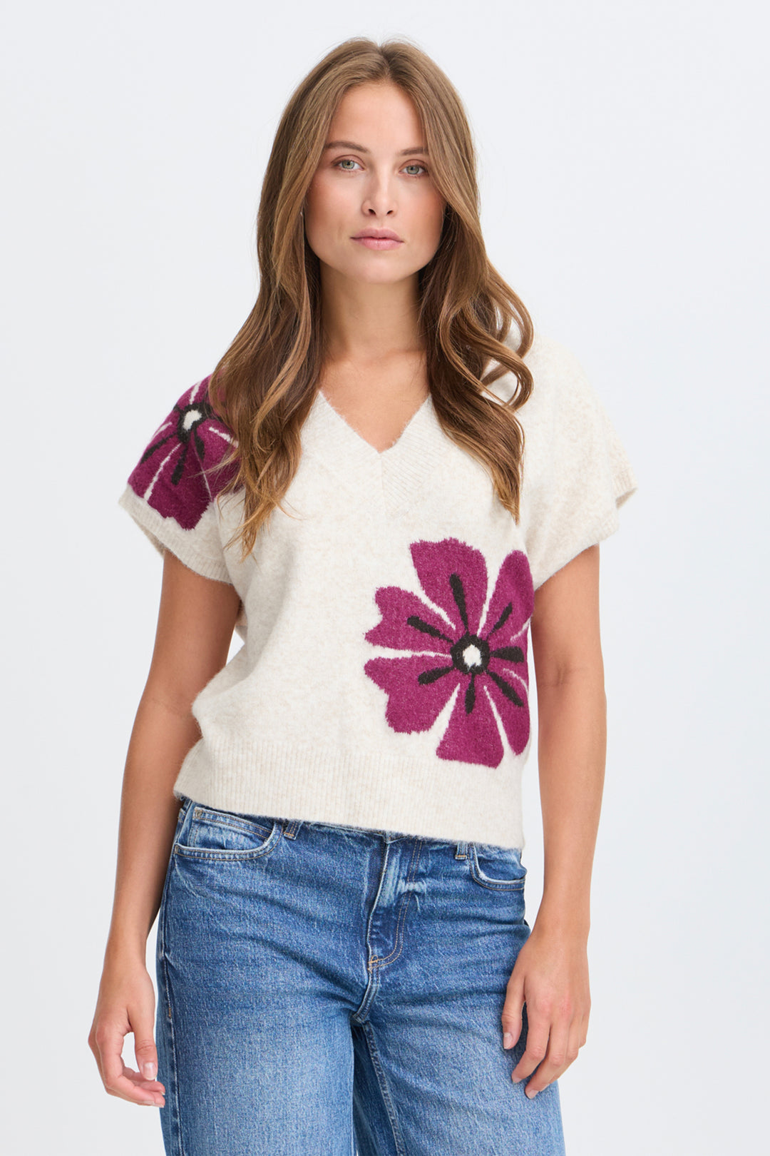 A woman modeling a cream-colored knit sweater with large magenta and purple flower motifs on the front and sleeves, paired with blue jeans against a light gray studio background.