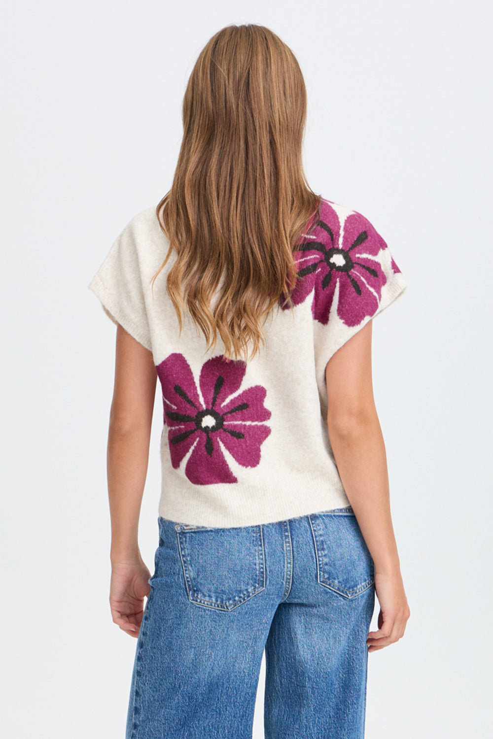 A woman viewed from the back wearing a cream knit top with large magenta floral motifs and blue jeans, against a plain light gray studio background.