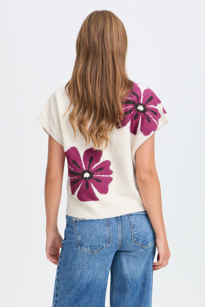 A woman viewed from the back wearing a cream knit top with large magenta floral motifs and blue jeans, against a plain light gray studio background.