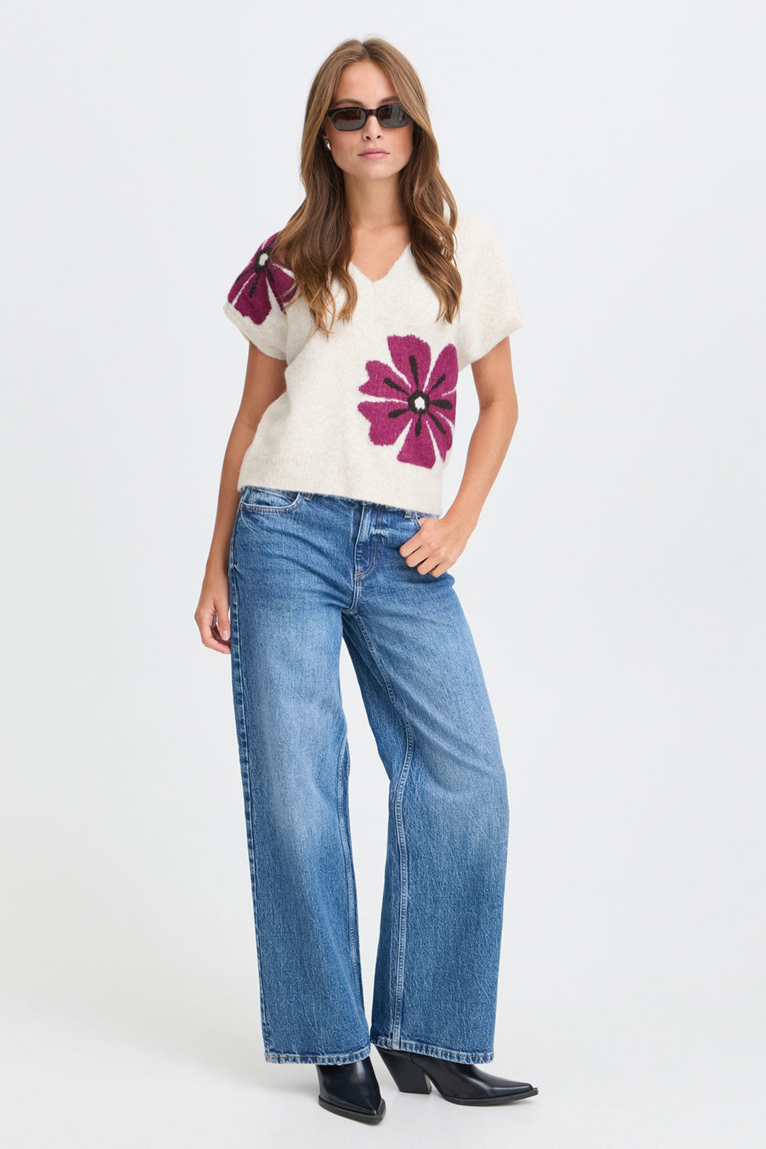 A woman modeling a cream knit top with large magenta flower appliqués on the front and sleeves, paired with wide-leg blue jeans and black pointed-toe heels, against a plain light gray studio background.