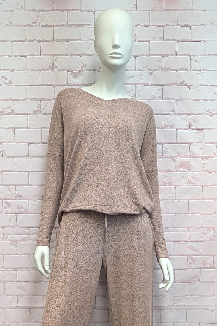 A mannequin wearing a light brown long-sleeve V-neck top and matching pants, standing against a light pink brick wall background.