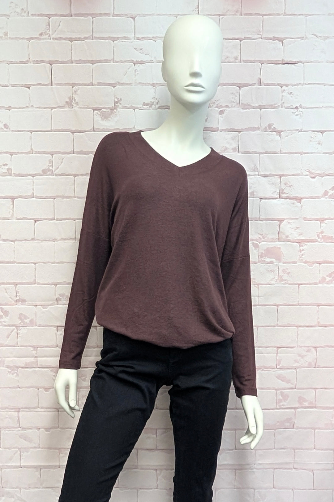 A mannequin wearing a dark brown long-sleeve V-neck top and black pants, standing against a light pink brick wall background.
