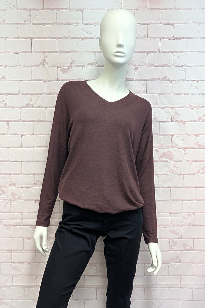 A mannequin wearing a dark brown long-sleeve V-neck top and black pants, standing against a light pink brick wall background.